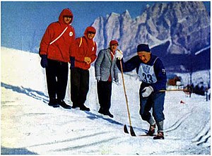 A man in a blue outfit with his competitor number «33» pinned to it skiing along a course in front of three other men.
